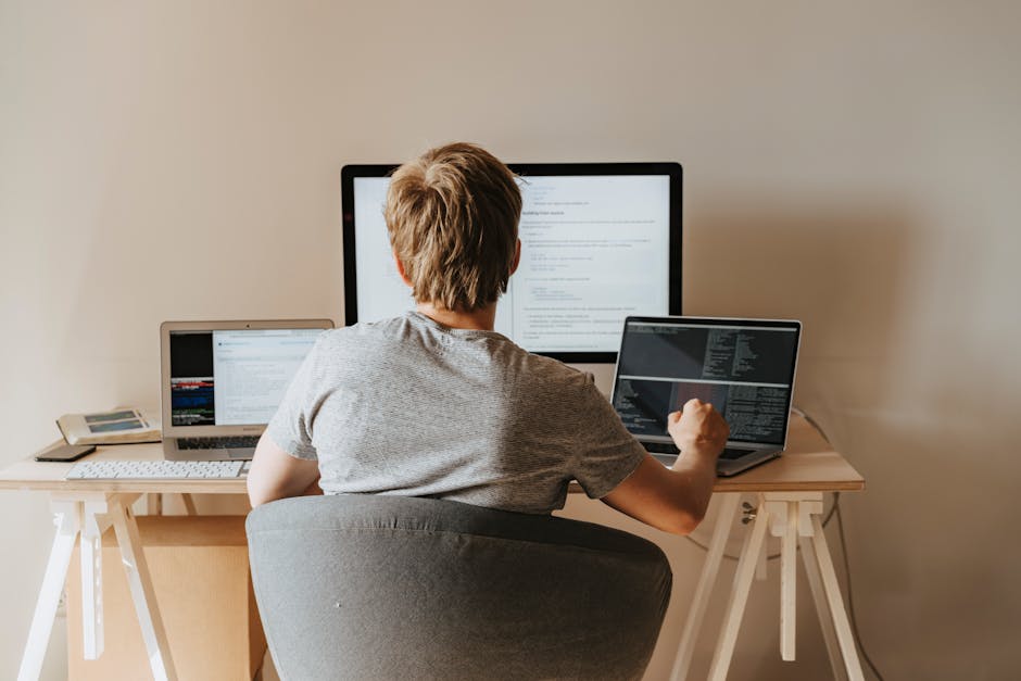 A young man with light brown hair, wearing a gray t-shirt, is seated at a wooden desk with a light-colored surface supported by white trestle legs. He is working on a desktop computer with a large mon