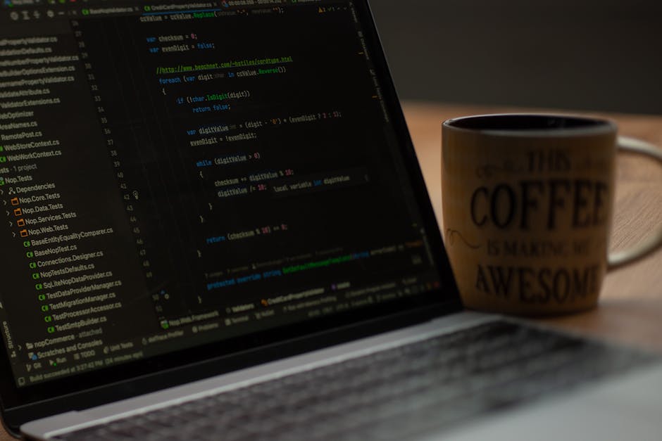 Close-up image of a laptop screen displaying lines of code in various colors, positioned on a wooden desk. Next to the laptop is a beige ceramic mug with black text that reads 'This coffee is making m